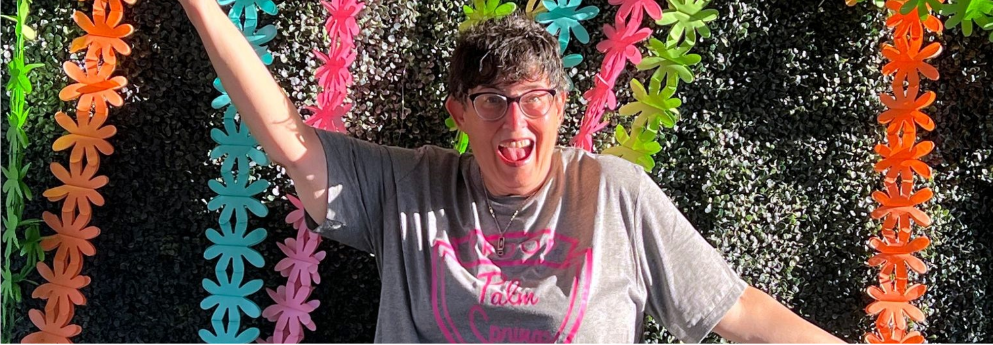 Stacy Gelman standing in front of a colorful wall smiling with her hands spread out wide
