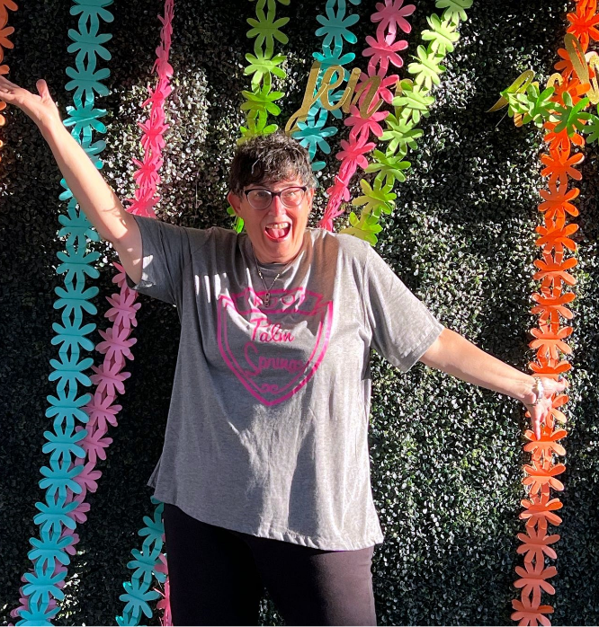 Stacy Gelman standing in front of a colorful wall smiling with her hands spread out wide